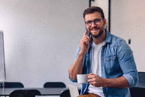 Handsome bearded man leaning on office desk, drinking coffee and talking on the phone.