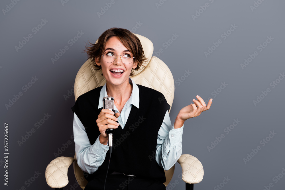 Photo of clever woman wear formalwear in glasses sit in armchair talk in mic look at offer empty space isolated on gray color background Photo of clever woman wear formalwear in glasses sit in armchair talk in mic look at offer empty space isolated on gray color background