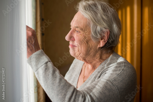 An elderly woman looking out a sunlit window with a reflective expression. photo