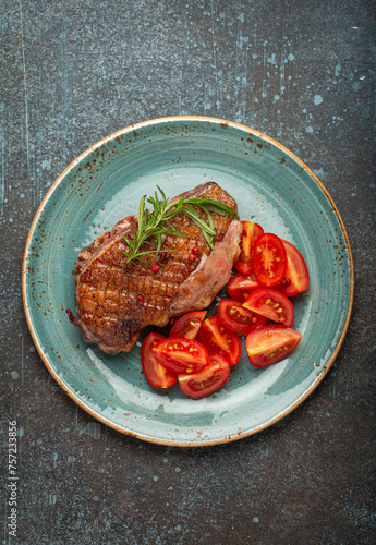 Delicious roasted duck breast fillet with golden crispy skin, with pepper and rosemary, top view on ceramic blue plate served with cherry tomatoes salad, rustic concrete rustic background