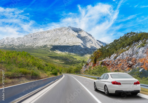 A white car drives along the highway against the backdrop of rocky mountains on a sunny day. © Denis Rozhnovsky