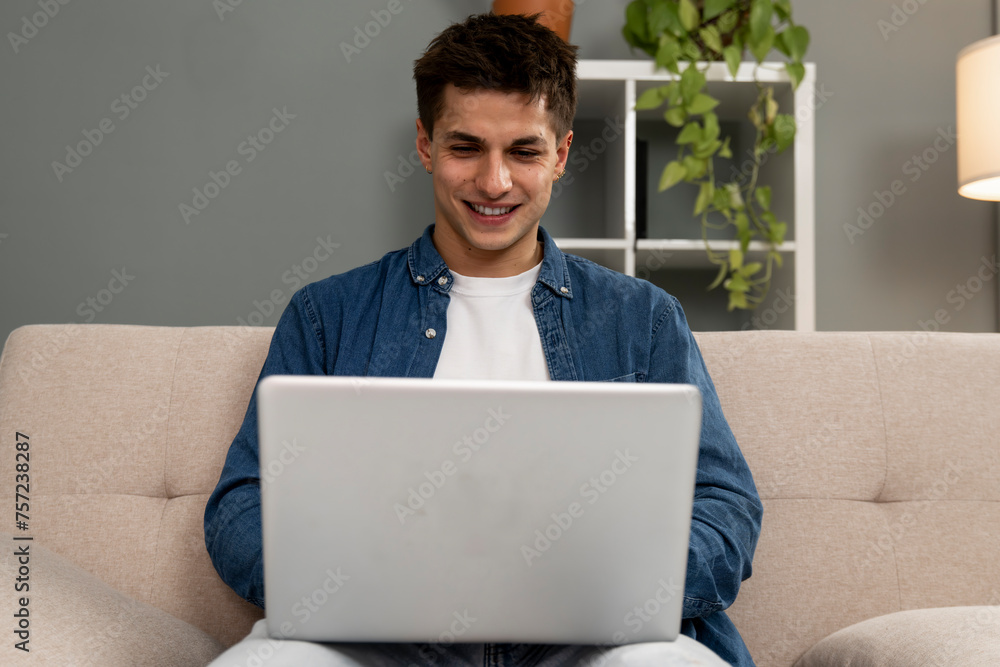 Handsome relaxed male student working with laptop while sitting on the couch. Smiling young carefree man using computer at home. Handsome relaxed male student working with laptop while sitting on the couch. Smiling young carefree man using computer at home.