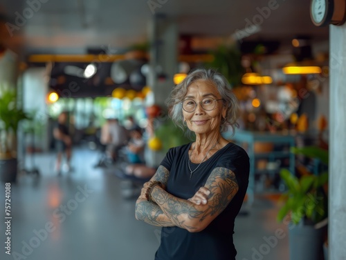 A happy old wrinkly elderly woman who is fitness instructor with visible tattoos covering muscled arms, with glassess is standing in the middle of the gym in sporstwear