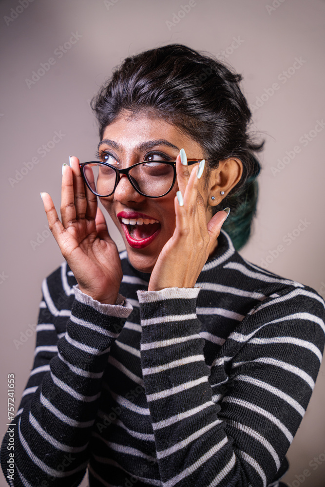 A woman with glasses and a red lip is smiling and making a funny face. She is wearing a striped shirt and has her hands on her face
