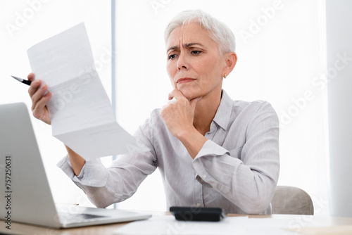 Confused Caucasian mature businesswoman paying bills using laptop at office. Middle-aged manager CEO HR analyzing reading bank statement, mortgage document, formal letter