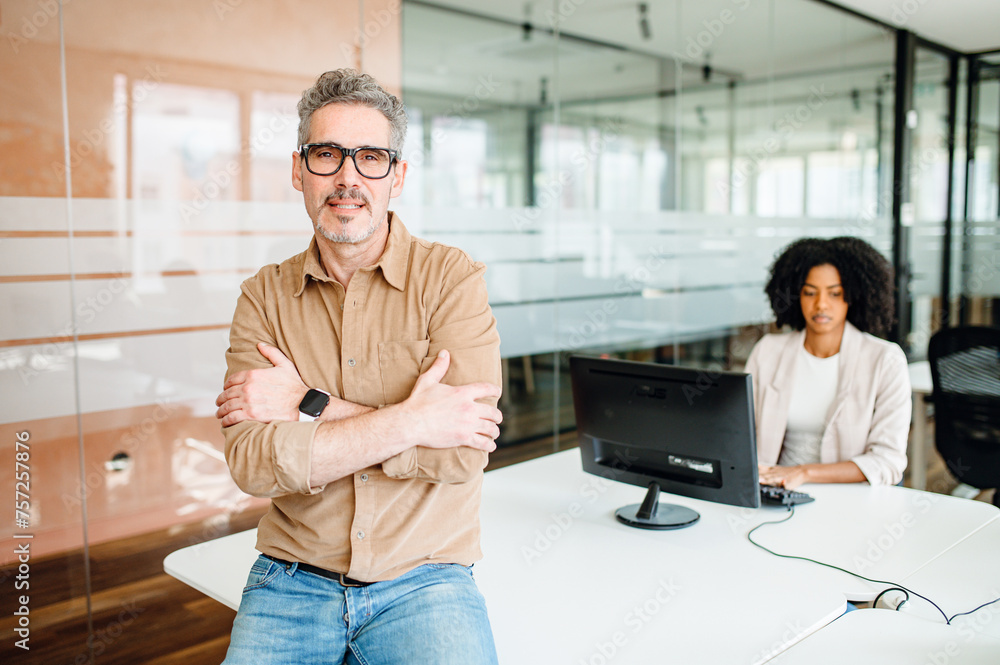 Senior leader stands with arms crossed in front of glass partition, a colleague working in the background, suggesting a supportive yet focused work environment that values leadership and concentration Senior leader stands with arms crossed in front of glass partition, a colleague working in the background, suggesting a supportive yet focused work environment that values leadership and concentration