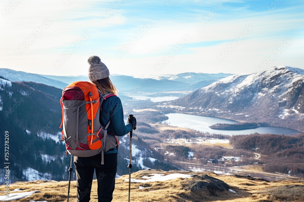 A hiker woman with a large backpack, hat and jacket stands a top a hill, gazing at a distant fjord and snowy mountains. Concept solo travelling, outdoor activities, adventure tourism.