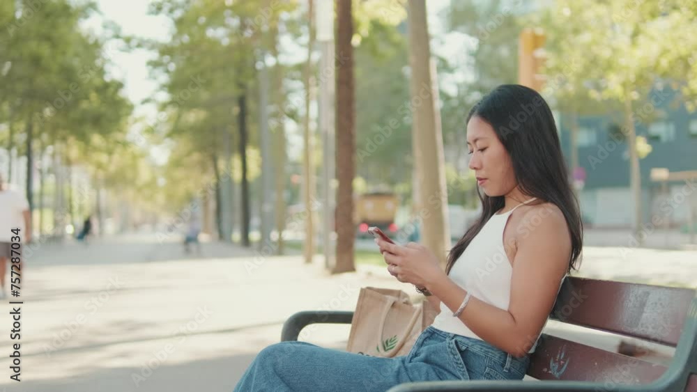 Young woman sitting on park bench typing message on mobile phone Young woman sitting on park bench typing message on mobile phone