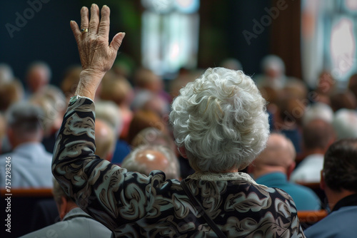 senior woman with hand up to talk at local town hall meeting, from back view
