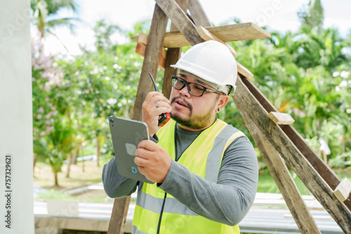 Male construction engineer uses walkie-talkie communicate with other builder in same construction area, communicate verbally tell construction worker on roof house needs so that they can follow plans. photo