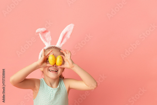 Portrait of cute smiling girl with Bunny ears and yellow Easter eggs, closes eyes with testicles, isolated on pink background. Happy Easter. Copy space