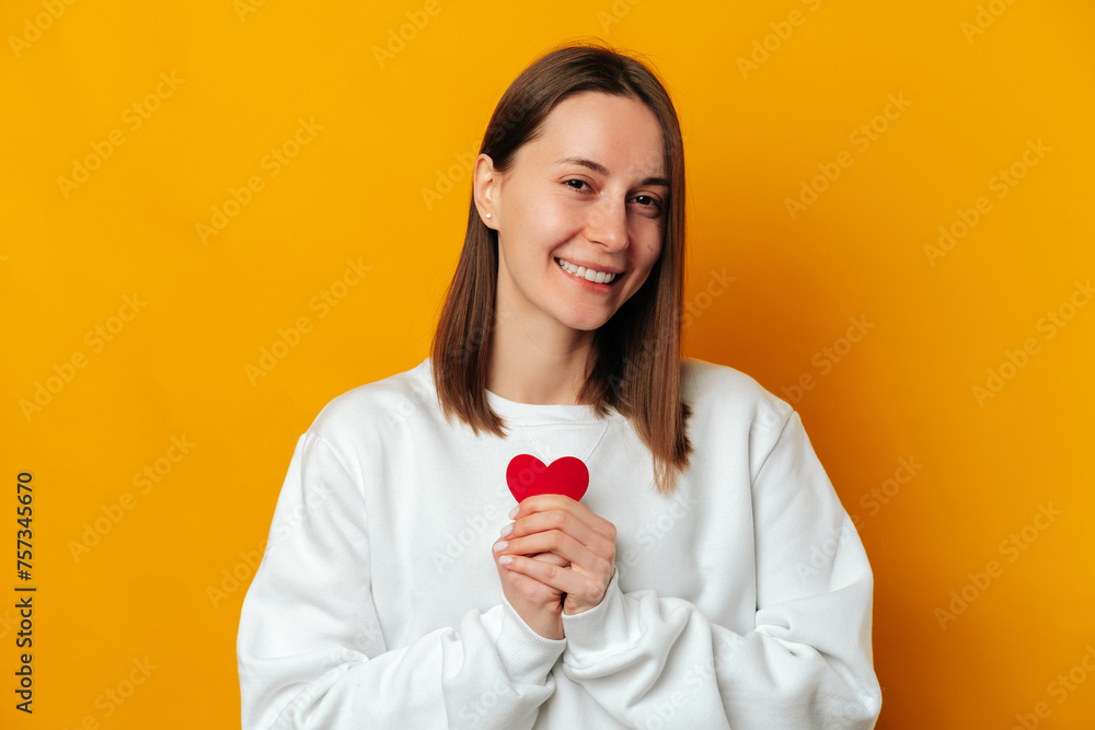 Cute young smiling woman wearing white is holding a red heart at her chest. Studio shot over yellow background. Cute young smiling woman wearing white is holding a red heart at her chest. Studio shot over yellow background.