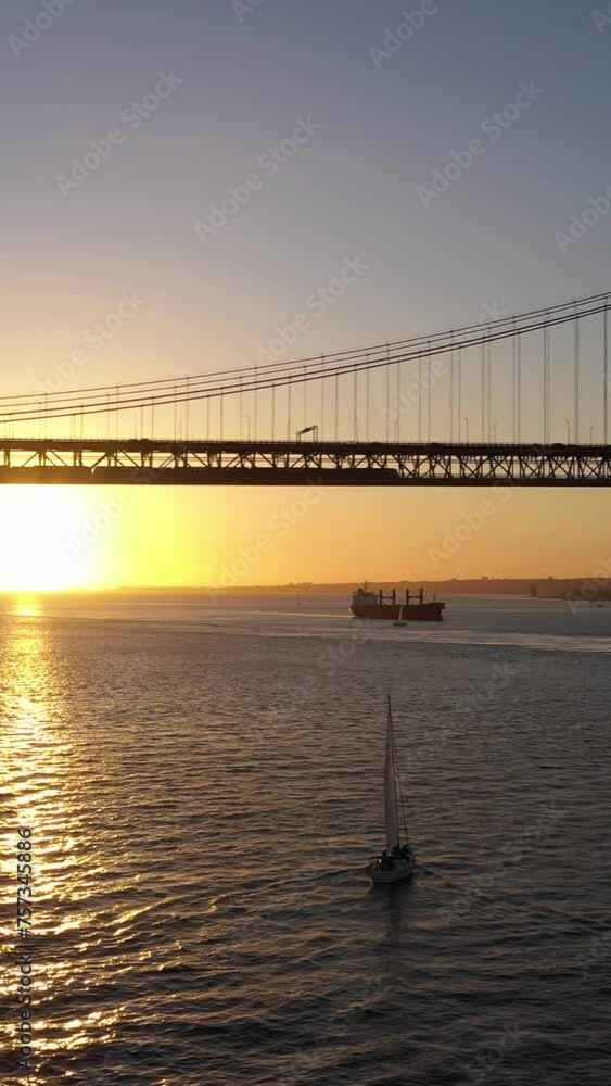 Silhouette of Ponte 25 de Abril Suspension Bridge at Sunset and Sailing Yacht. 25th April Bridge. Portugal. Aerial View. Drone Moves Forward and Upwards. Vertical Video Silhouette of Ponte 25 de Abril Suspension Bridge at Sunset and Sailing Yacht. 25th April Bridge. Portugal. Aerial View. Drone Moves Forward and Upwards. Vertical Video
