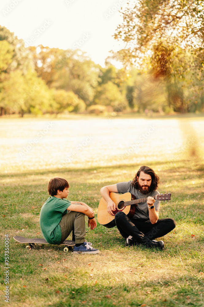 Vertical outdoors shot of a bearded man teaching a boy how to play guitar while sitting on grass in a park. Vertical outdoors shot of a bearded man teaching a boy how to play guitar while sitting on grass in a park.