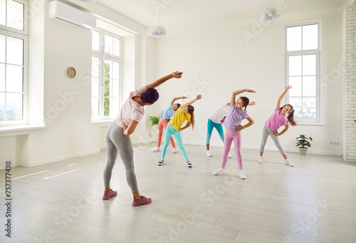 Female young choreographer showing her students girls stretching exercises in dance studio. Happy kids doing dance workout. Children training in choreography class, kids sport concept.