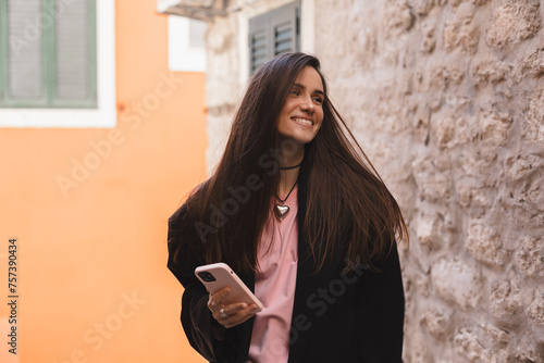 Photo of charming pretty young lady hold modern gadget smiling outside urban city street. Pensive brunette woman student freelancer wear black jacket walking in old town, hold phone in hand.