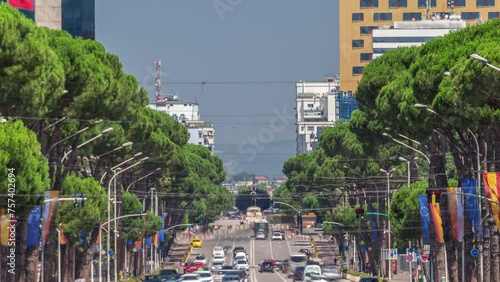 Traffic on the Deshmoret e Kombit Boulevard in Tirana timelapse. Main street in Albanian capital surrounded by green trees close up view. Cars moving and stops on intersections with a traffic light photo