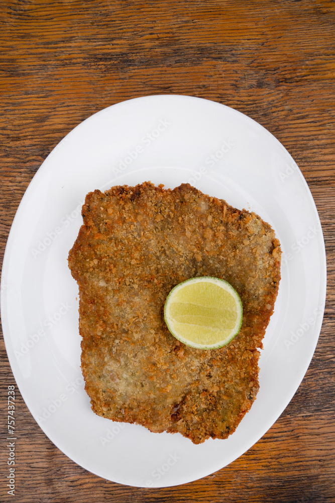 Argentine dish. Top view of milanesa, breaded fried steak, with a slice of lemon, in a white dish with a wooden background. Argentine dish. Top view of milanesa, breaded fried steak, with a slice of lemon, in a white dish with a wooden background.