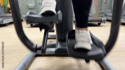 Close-up video: moving legs in white sports shoes on the exercise machine. Mature woman training on an elliptical trainer. Slow motion in the gym. The concept of health and sports in middle age.