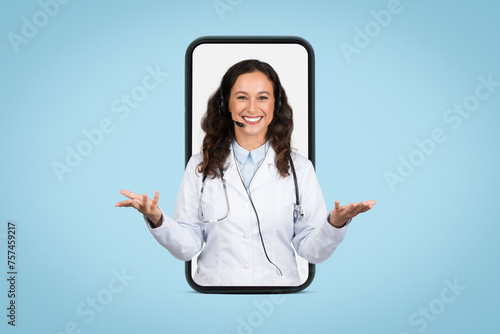Friendly european female doctor with headset in giant smartphone screen gesturing and smiling at camera