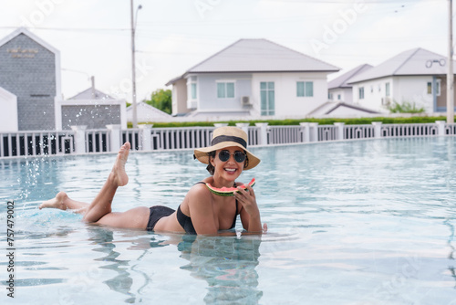 Beautiful Spanish woman lying on edge of swimming pool Wear sunglasses, a sun hat and a swimsuit. Woman lies down and eats juicy red watermelon comfortably was wet because had just finished swimming. © Ekkasit A Siam