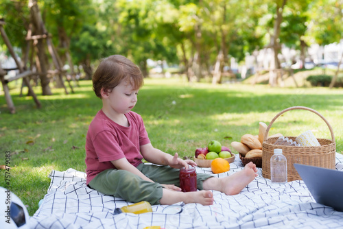 Adorable Caucasian boy sitting on grass in village park, hungry, dipping finger into jam jar and sucking it with gusto, hunger after playing until tired, family vacation Concept #757479083