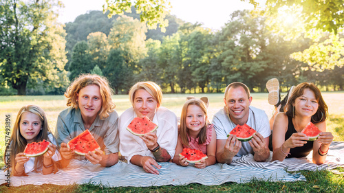 Big family lying on the picnic blanket in city park under Linden tree during weekend Sunday sunny day, smiling, laughing at camera and eating watermelon. Family values and outdoor activities concept.