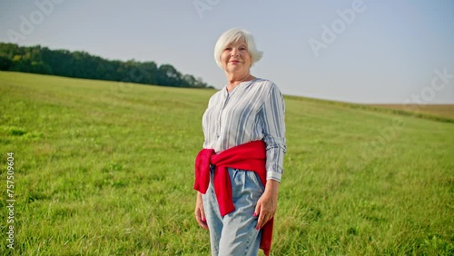 Zooming in on older Caucasian woman with white hair. Female standing on large hill with short grass and looking directly at camera. Wonderful horizon in background. Positive attitude.