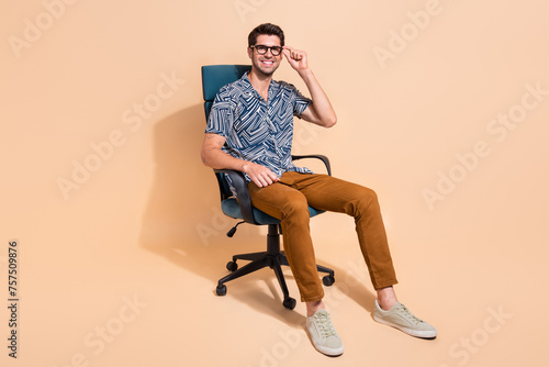 Full length photo of handsome businessman touching specs sitting comfortable boss chair enjoying office isolated on beige color background