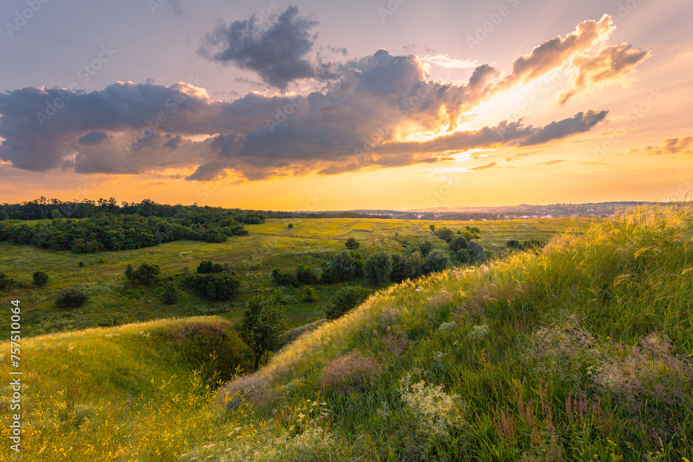 Obraz premium The majestic dusk scene with lush grass and wildflowers covered hills in front of beautiful sunset sky with clouds. Summer nature background.