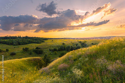 The majestic dusk scene with lush grass and wildflowers covered hills in front of beautiful sunset sky with clouds. Summer nature background.