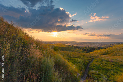 The majestic dusk scene with motley high grass and wildflowers covered hills in front of a beautiful sunset sky with clouds. Summer nature background. © stone36