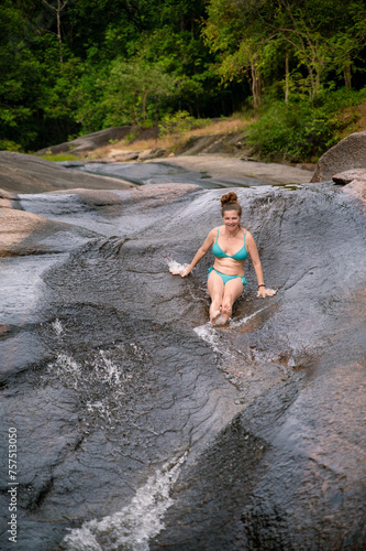 A woman glides with joy down a smooth, natural rock slide in the verdant forests of Langkawi, embracing the fun of nature