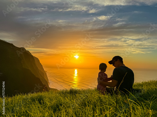 Father playing with toddler on meadow with breathtaking sunset at viewing point Miradouro do Ponta da Ladeira  Madeira island  Portugal  Europe. Panoramic view of majestic coastline Atlantic Ocean