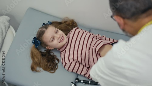 Over-the-shoulder shot. A little girl patient lies on her back on the medical examination couch and laughs while her abdomen is examined by a pediatrician. A male doctor palpation a child's abdomen photo