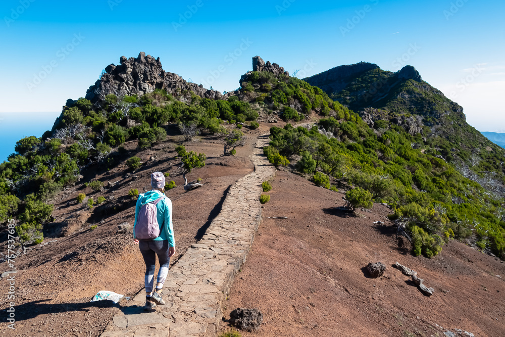 Hiker woman on scenic paved hiking trail winding up to mountain peak Pico Ruivo, Madeira island, Portugal, Europe. Path on rugged terrain of unique rock formations and deserted shrub land. Wanderlust