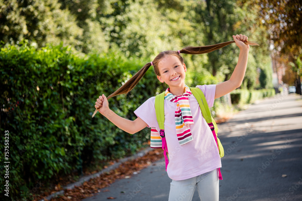 Photo of positive good mood little girl dressed tied jumper rucksack holding ponytails having fun outdoors urban city park