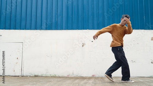 Hispanic man stretch arms and dance street dancing in front of wall. Motion shot of stylish dancer or choreographer in casual outfit practicing dancing in hip hop style. Outdoor sport 2024. Endeavor.