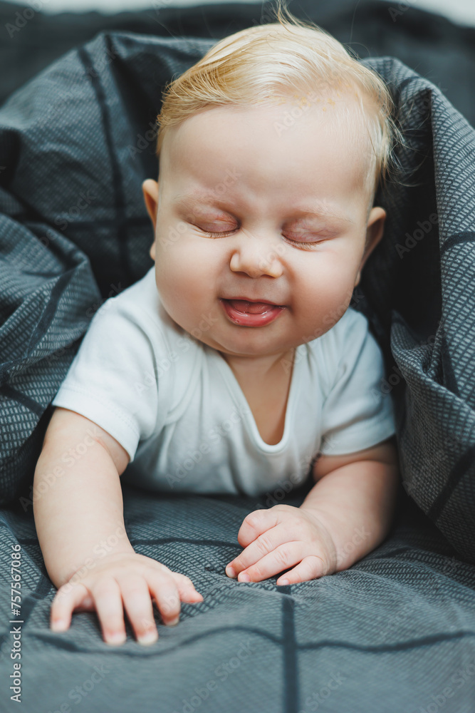 A cheerful baby boy with beautiful big eyes is lying on the bed at home in a white bodysuit on gray bedding. The child is lying in bed at home A cheerful baby boy with beautiful big eyes is lying on the bed at home in a white bodysuit on gray bedding. The child is lying in bed at home