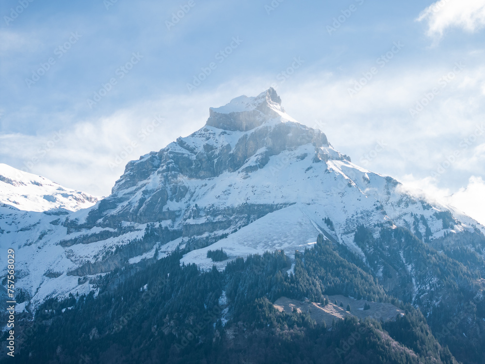 Early morning or late afternoon light bathes a snowy Engelberg mountain peak, with rocky outcrops and forested slopes beneath a pale blue sky with wispy clouds.