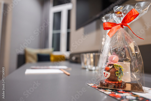 Billede på lærred Closeup of a whimsical chocolate gift wrapped in cellophane with a red ribbon, featuring colorful details on a patterned base, set against a modern room's blurred interior in Engelberg