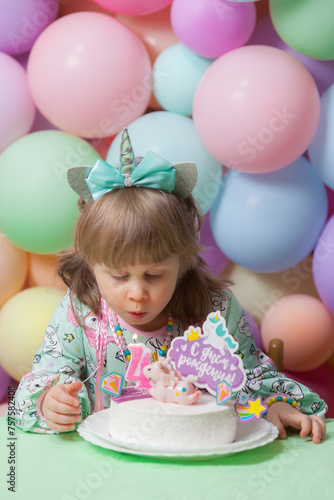 A little girl in a unicorn dress and hairband is blowing out a birthday candle on her cake isolated on a pastel latex balloons background. Vertical image.
