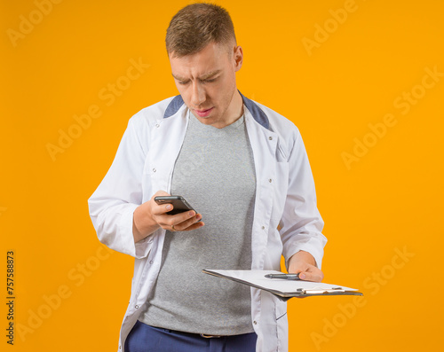 A medical professional distracted by his phone while holding a clipboard, against an orange backdrop.