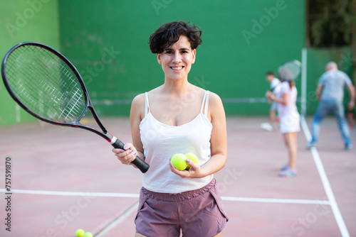 Portrait of positive latin woman frontenis player standing on outdoor fronton, holding racket and tennis ball, smiling and looking at camera. © JackF