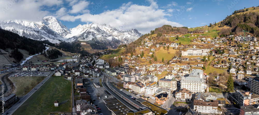 Panoramic view of Engelberg, Switzerland, showcasing its alpine village, green fields, accessible roads, and snowcapped mountains, highlighting the area's natural beauty and recreational appeal.