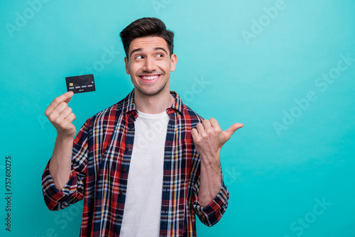 Photo of cheerful dreamy man dressed plaid shirt rising bank card pointing thumb empty space isolated turquoise blue color background © deagreez