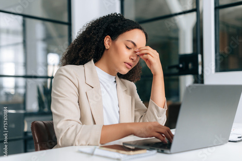 Exhausted upset brazilian or hispanic curly business woman, company employee, working on a laptop in modern office, has a headache, migraine, massages nosebridge with eyes closed, feels overworked photo