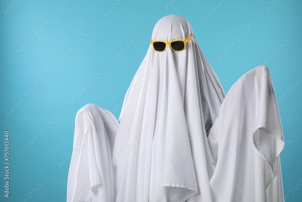 Stylish ghost. Person covered with white sheet in sunglasses on light blue background