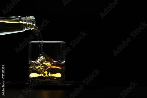 Pouring whiskey from bottle into glass with ice cubes at table against black background, space for text