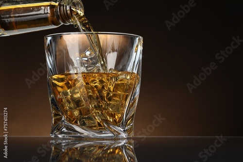 Pouring whiskey from bottle into glass with ice cubes at table against brown background, space for text